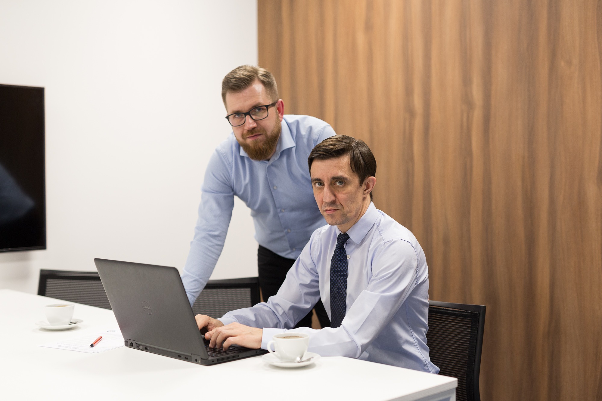 Dwóch mężczyzn w biurze przy białym stole z laptopem i filiżankami kawy, jeden siedzi i pisze na laptopie, drugi stoi za nim i patrzy w obiektyw.