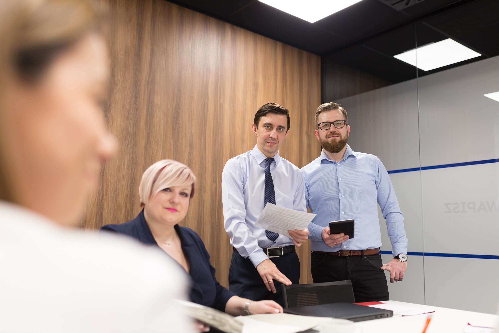 Spotkanie biznesowe w biurze w Bełchatowie: kobieta siedzi przy stole, dwóch mężczyzn stoi, jeden trzyma dokumenty, drugi tablet, w tle drewniana ściana i szklana ściana z napisem.