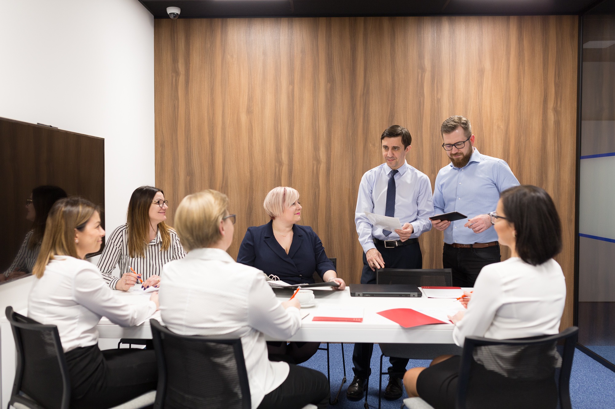 Zespół osób w sali konferencyjnej, dwóch mężczyzn prezentuje dane na tablecie i papierze, kobiety przy stole notują, nowoczesne wnętrze z drewnianą ścianą.