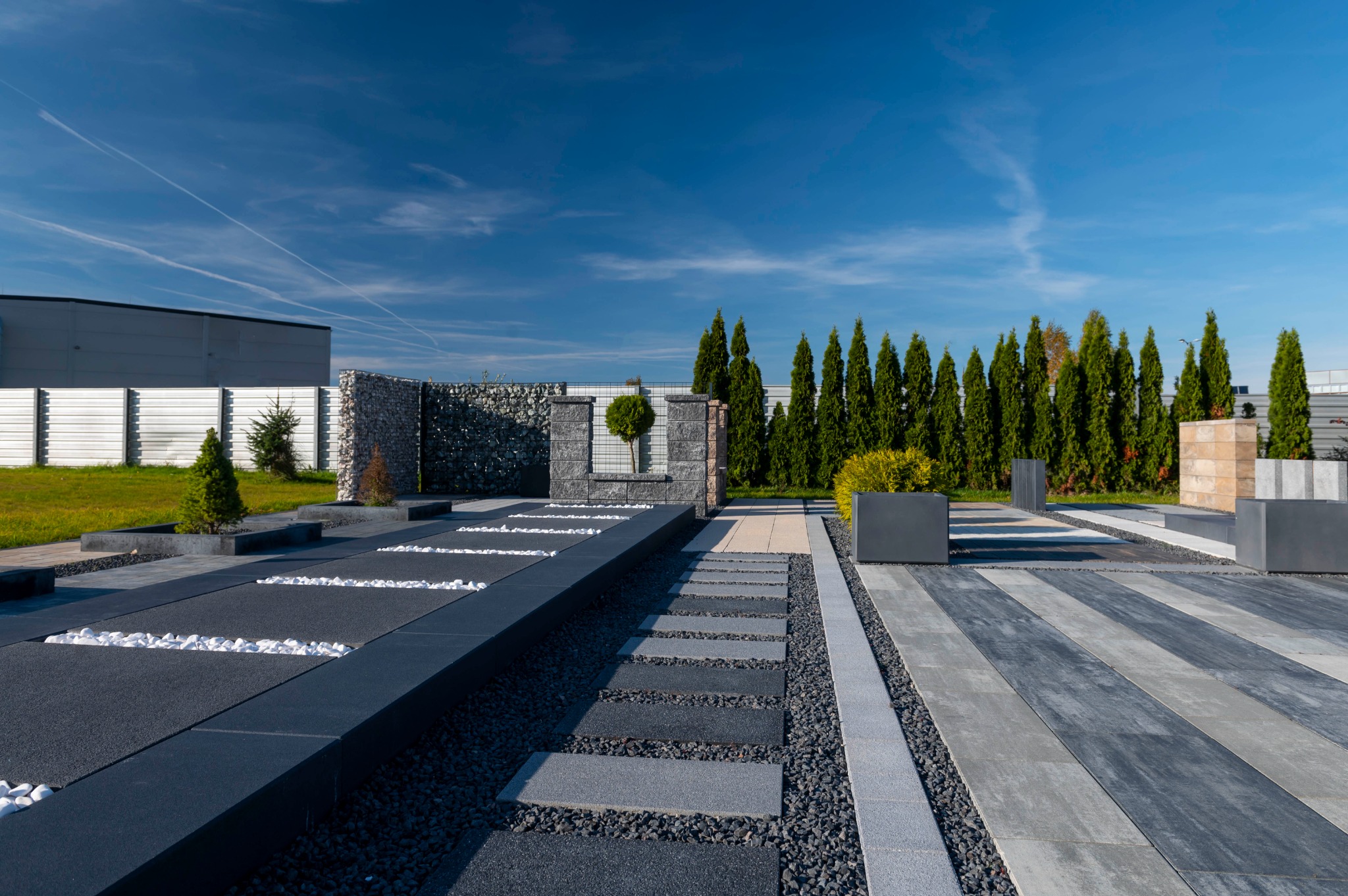 Wzorcowa ekspozycja różnorodnych nawierzchni: szare i czarne płyty chodnikowe, ścieżki z drobnych kamyczków, donice z zielenią i gabionowe ogrodzenie w tle, na tle błękitnego nieba.