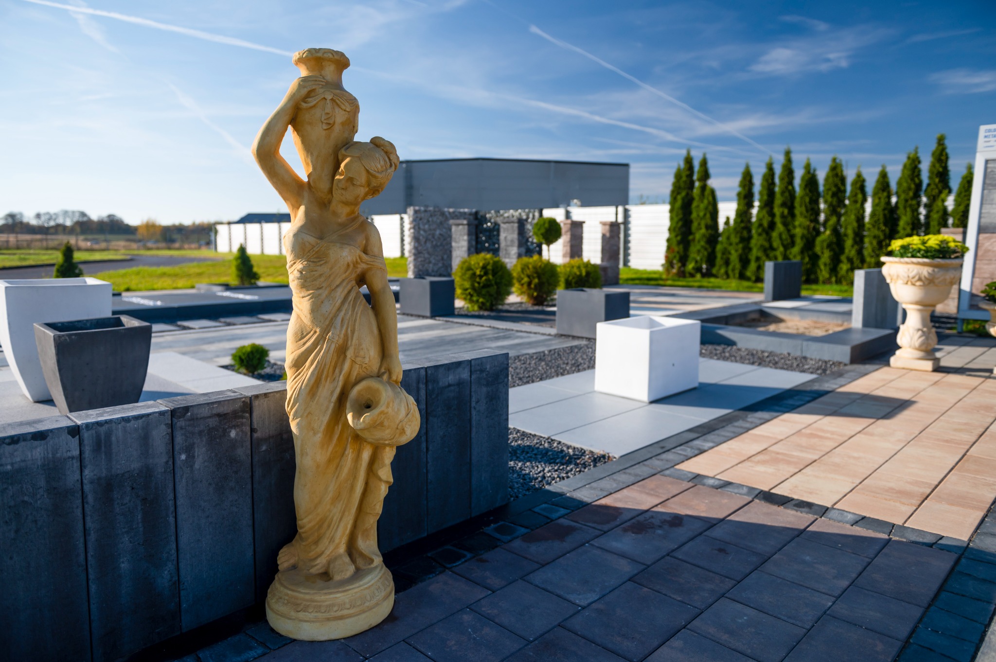 Ekspozycja zewnętrzna z rzeźbą kobiety niosącej dzban, obok donice i fragmenty nawierzchni z różnokolorowej kostki brukowej w słoneczny dzień.