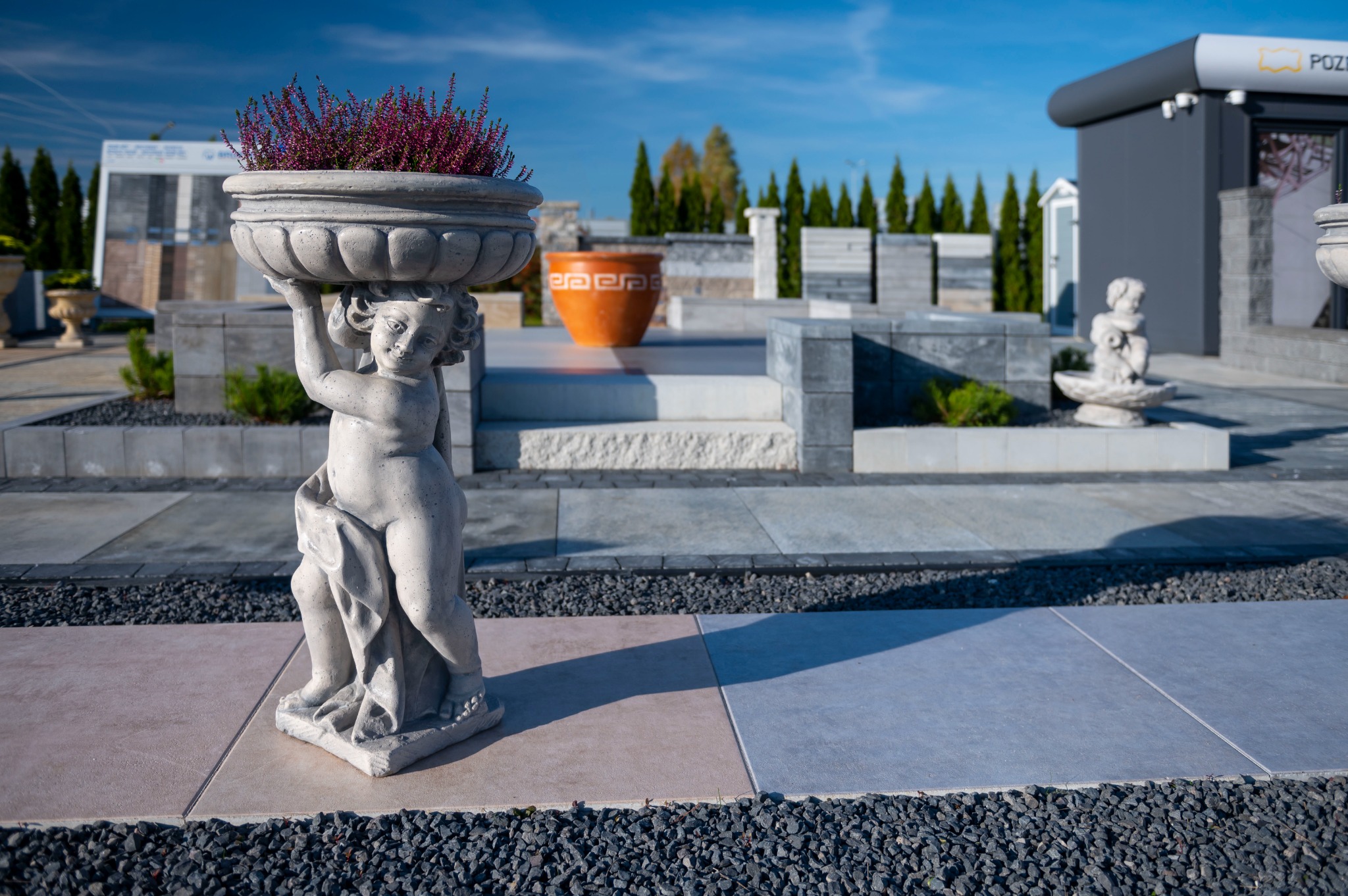 Figurka aniołka podtrzymująca betonową donicę z wrzosem na tle ekspozycji nawierzchni z płyt betonowych i aranżacji ogrodowych.