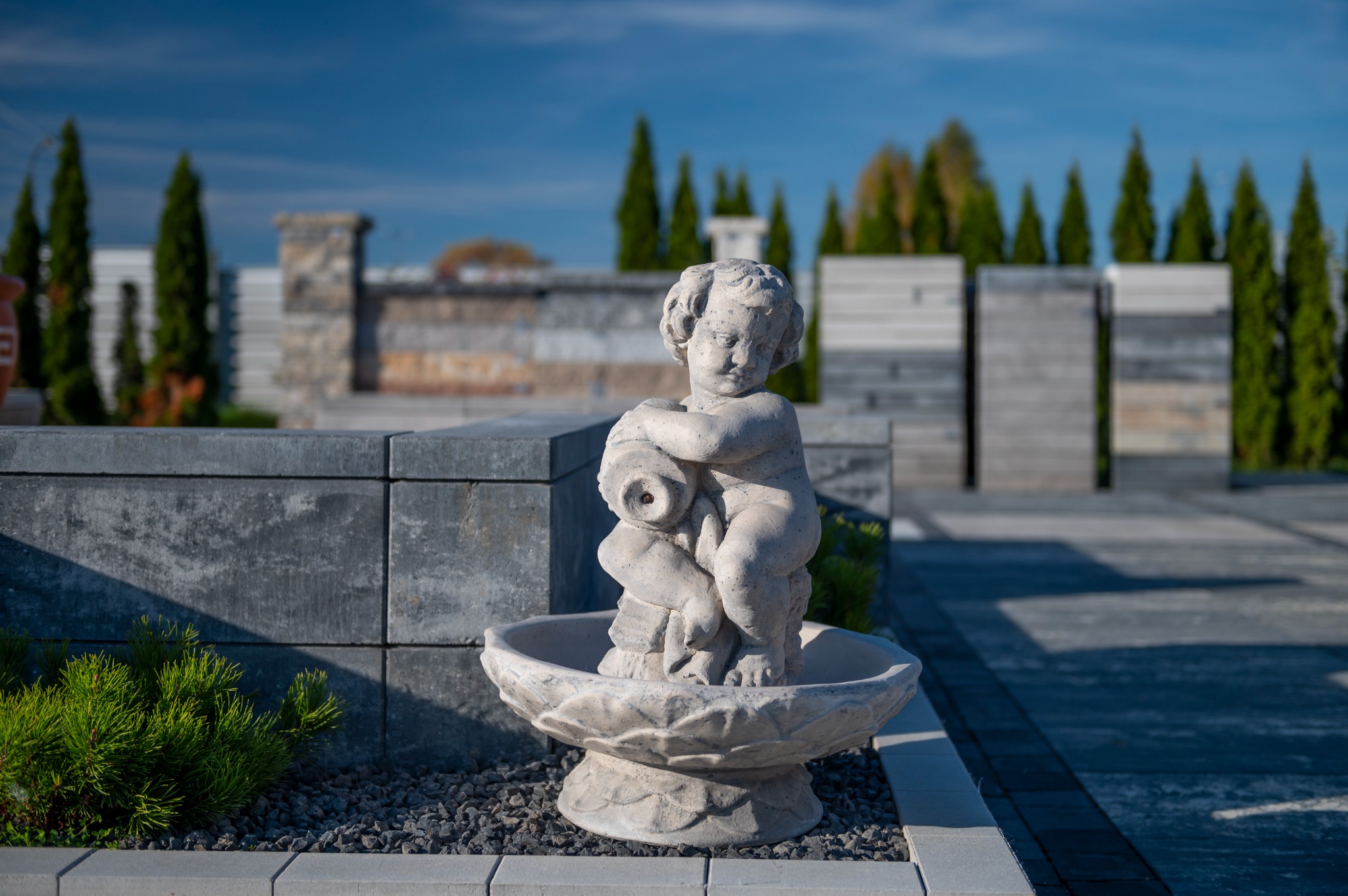 Fontanna ogrodowa z figurką cherubina na tle nowoczesnego ogrodzenia z prefabrykatów betonowych i strzyżonych iglaków.