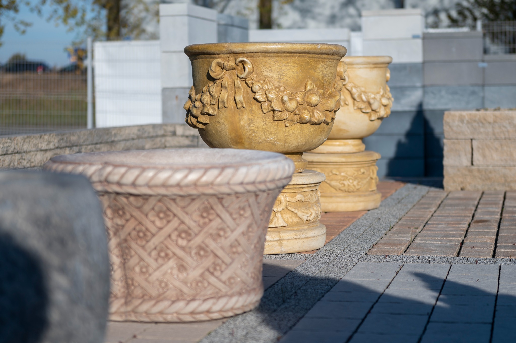 Dekoracyjne, beżowe donice betonowe o różnych wzorach, ustawione na wybrukowanym chodniku z kostki, z paletą betonowych elementów w tle.