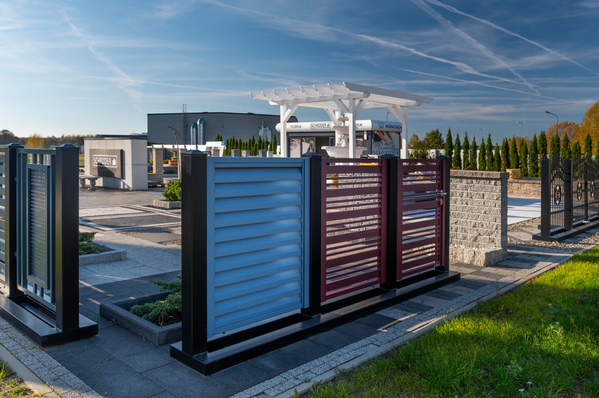 Ekspozycja różnych typów ogrodzeń: panelowe w kolorze niebieskim i bordowym, metalowe kute i nowoczesne, na tle pawilonu ogrodowego i budynku firmy.