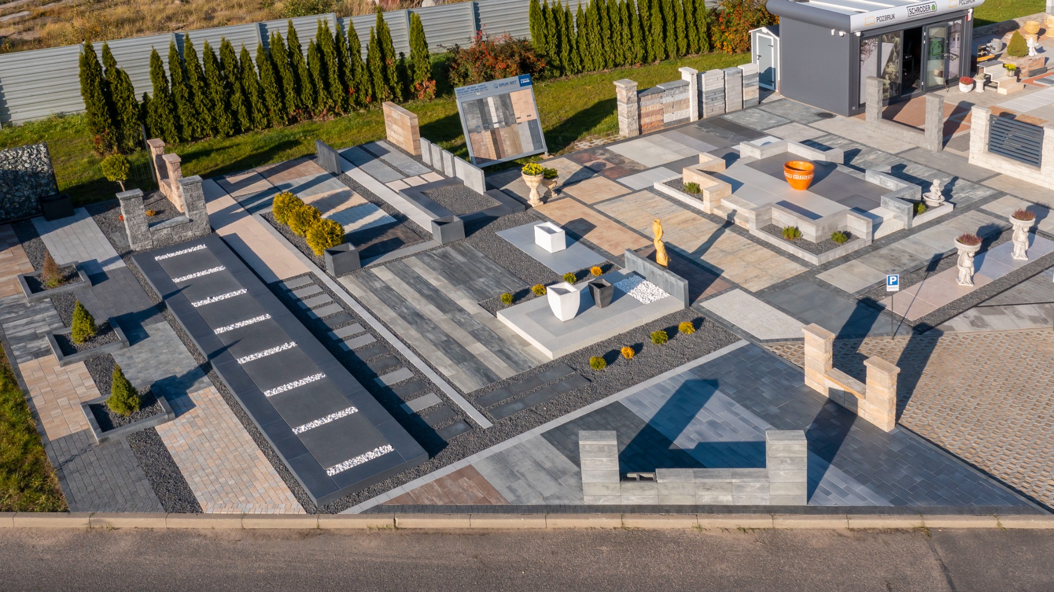 Ekspozycja różnorodnych nawierzchni z kostki brukowej i elementów betonowych na zewnątrz salonu sprzedaży, widok z góry, prezentacja aranżacji z roślinami ozdobnymi i elementami małej architektury.