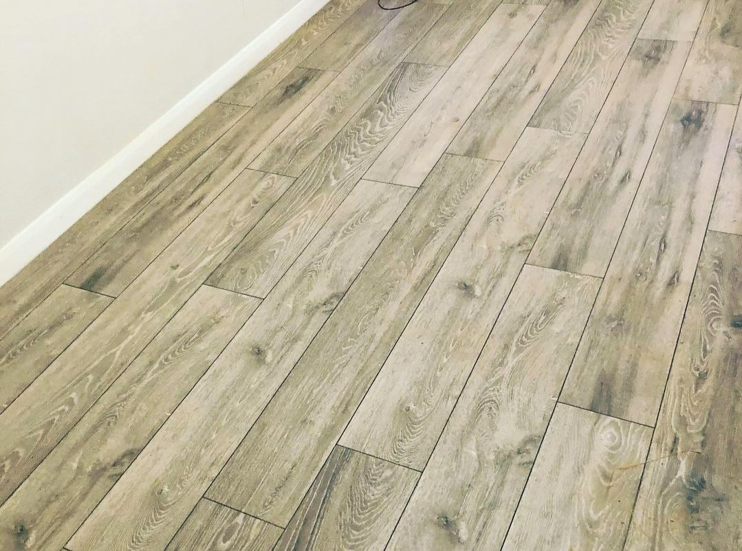 Ułożone panele podłogowe w odcieniach szarości, z widoczną strukturą drewna, ułożone diagonalnie względem ściany z białą listwą przypodłogową.