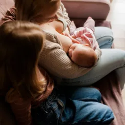 Mama karmiąca piersią noworodka w ramionach, obok siedzi starsza córka, ciepłe światło z okna.