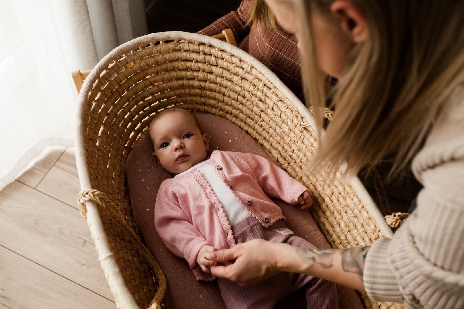 Noworodek w różowym ubranku leżący w wiklinowym koszu, trzymany za rękę przez osobę z tatuażem na przedramieniu.