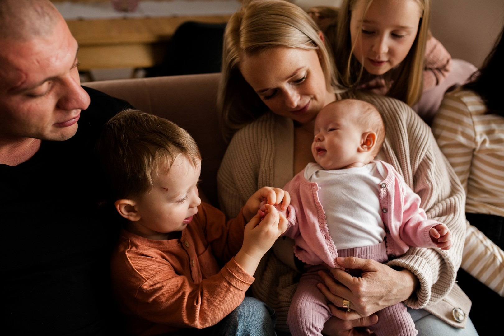 Mama trzyma na rękach niemowlę w różowym sweterku, obok siedzi mały chłopiec w pomarańczowej koszuli, a za nimi stoi starsza dziewczynka, wszyscy patrzą na dziecko.