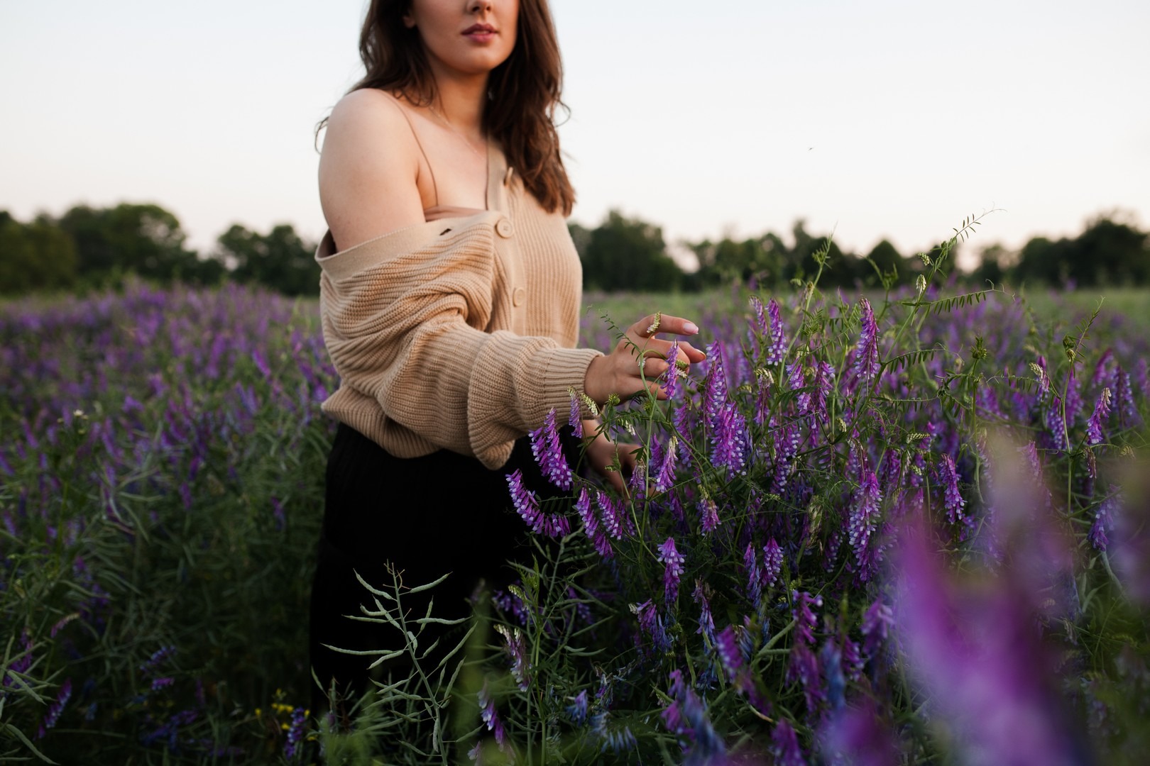 Kobieta w beżowym swetrze dotyka delikatnych, fioletowych kwiatów na rozległej łące w miękkim, naturalnym świetle.