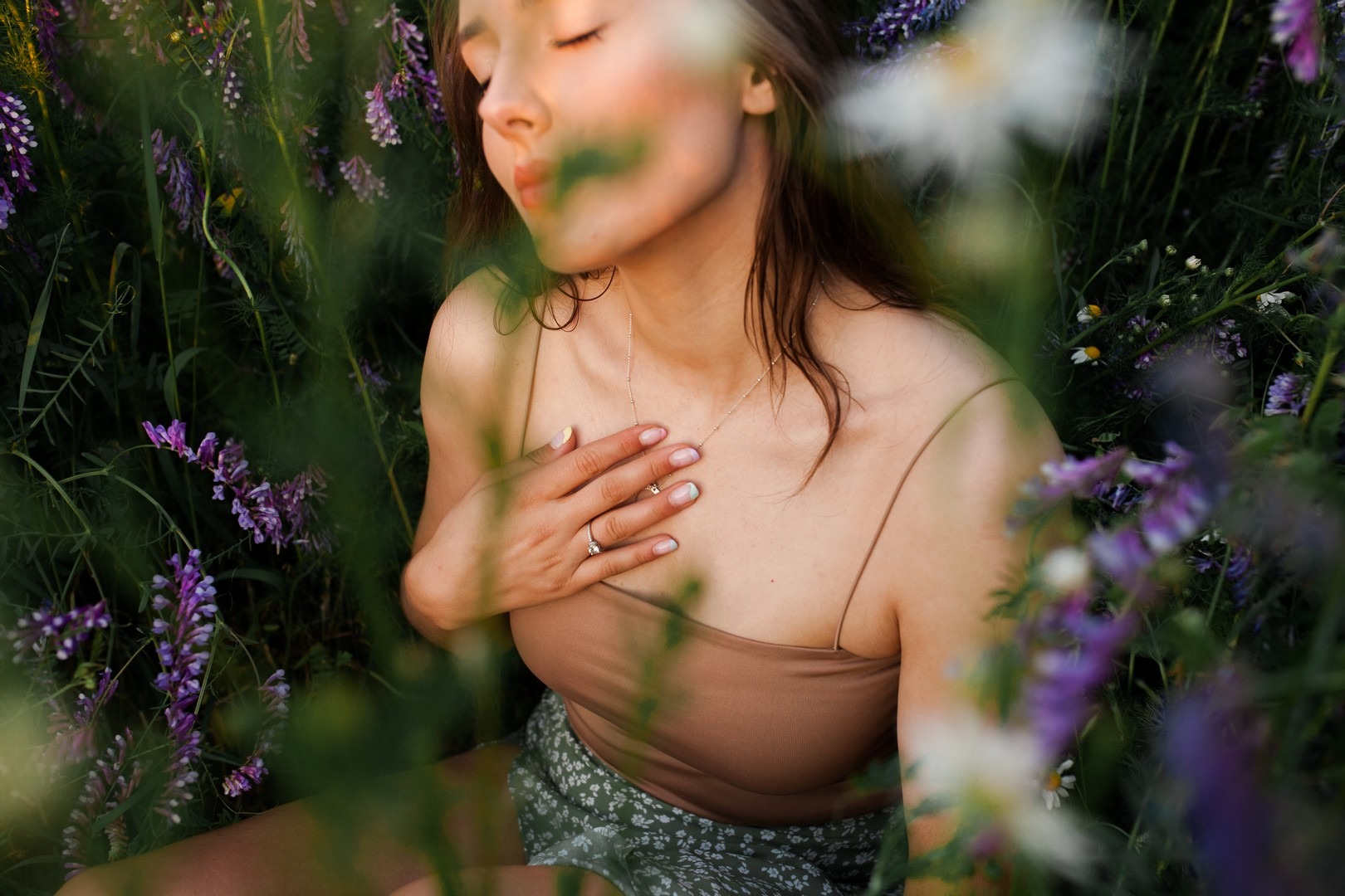 Młoda kobieta z zamkniętymi oczami, siedząca w kwitnącej łące, delikatnie dotyka dłonią dekoltu, na palcu pierścionek zaręczynowy, subtelny makijaż, pastelowe paznokcie.