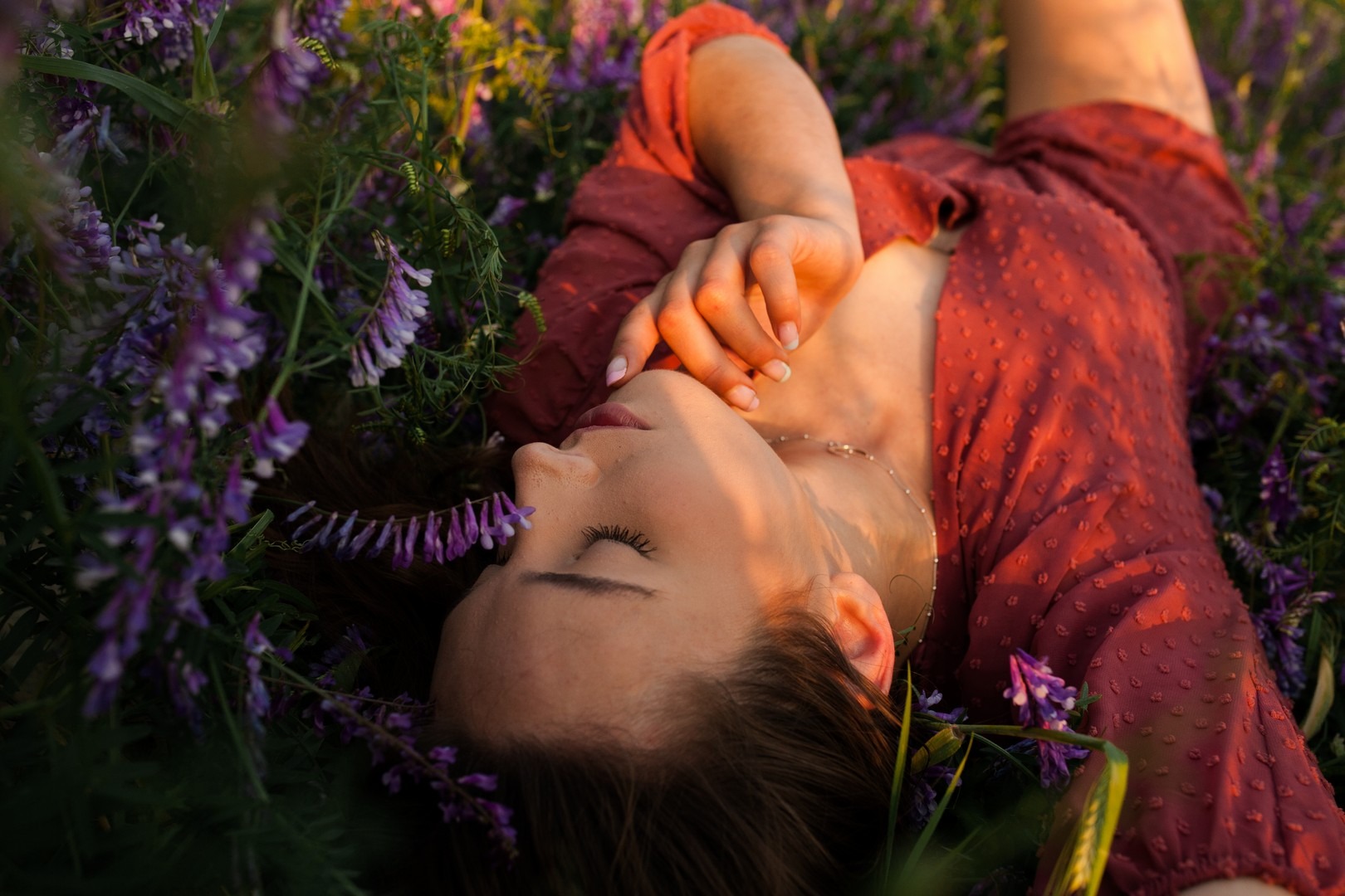 Portret kobiety w czerwonej sukience leżącej w kwitnącej łące, delikatnie dotykającej twarzy, z widocznym naszyjnikiem i manicure.