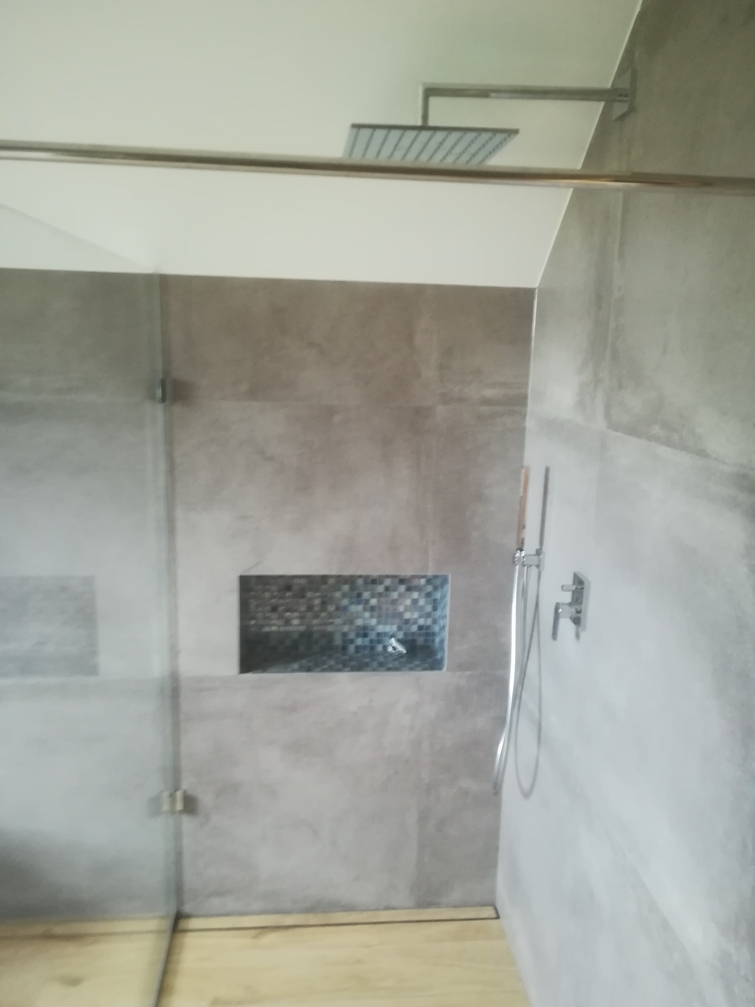 Nowoczesna kabina prysznicowa z szarą, betonową fakturą płytek, wnęką z mozaiką i deszczownicą zamontowaną na suficie, widoczna szklana ściana z metalowymi zawiasami.