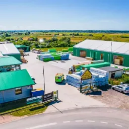 Widok z lotu ptaka na teren składu drewna z budynkami o zielonych dachach, zaparkowanymi samochodami i stertami drewna na zewnątrz oraz w magazynie.