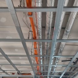 T&T Paweł Knutti - Sufit podwieszany z widocznymi kablami elektrycznymi w pomarańczowej osłonie, srebrnymi rurami wentylacyjnymi w izolacji oraz metalową konstrukcją szkieletową, w trakcie prac wykończeniowych.