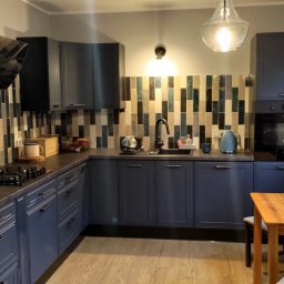 T&T Paweł Knutti - Niebieska kuchnia z mozaiką na ścianie, drewnianym stołem i krzesłami. Widoczny piekarnik, płyta gazowa i okno z grzejnikiem. Nowoczesny design.