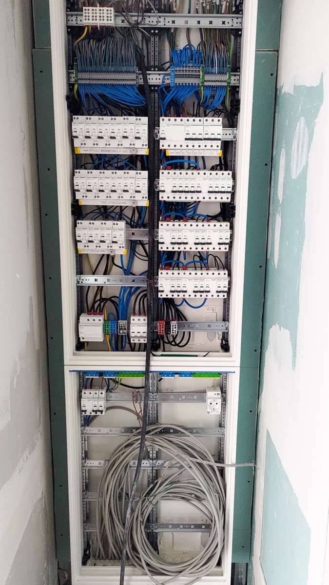 Rozdzielnica elektryczna w trakcie montażu, widoczne liczne przewody, zabezpieczenia i elementy montażowe w obudowie osadzonej w ścianie z płyt gipsowo-kartonowych.