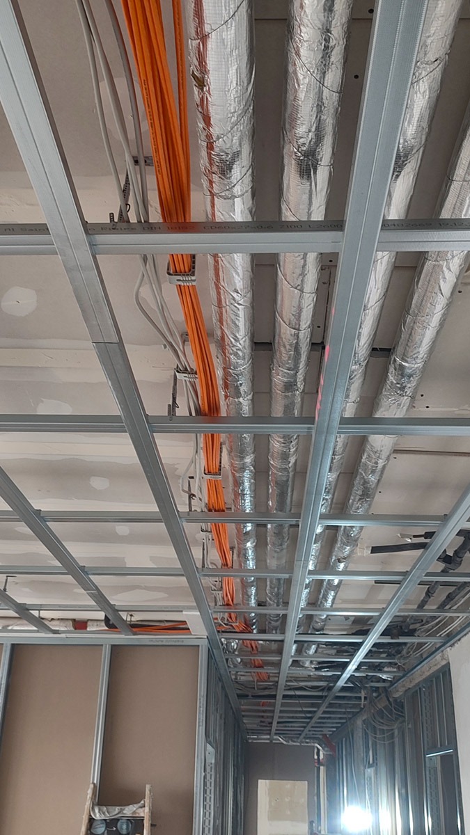 Sufit podwieszany z widocznymi kablami elektrycznymi w pomarańczowej osłonie, srebrnymi rurami wentylacyjnymi w izolacji oraz metalową konstrukcją szkieletową, w trakcie prac wykończeniowych.