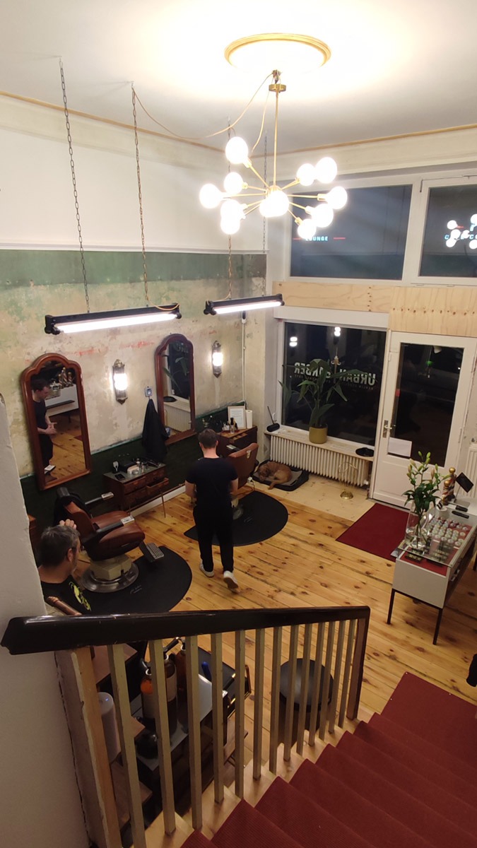 Widok z góry na wnętrze barber shopu z drewnianą podłogą, dwoma fotelami barberskimi, lustrami z oświetleniem, centralnym żyrandolem w stylu retro i widoczną instalacją elektryczną z kablami...