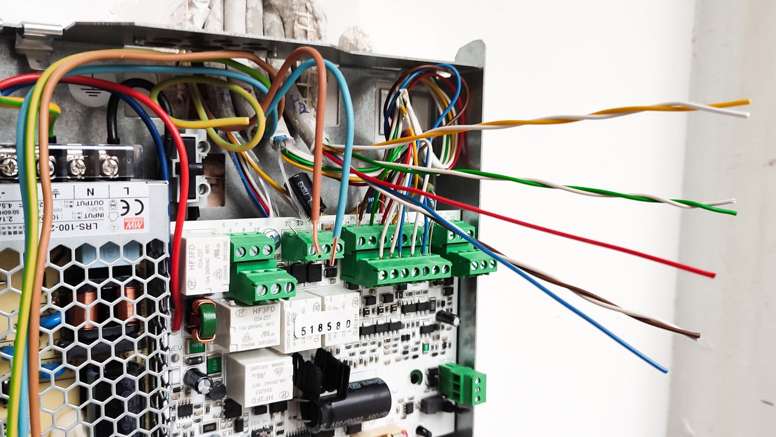 Otwarta skrzynka elektryczna z widocznymi przewodami o różnych kolorach podłączonymi do zielonych złączek na płytce drukowanej, zasilacz impulsowy LRS-100-2 oraz liczne komponenty elektroniczne...