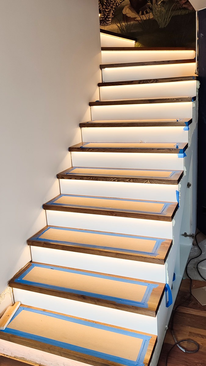 Schody z podświetleniem LED na każdym stopniu, zabezpieczone taśmą malarską, widoczne okablowanie, w tle ściana z tapetą.
