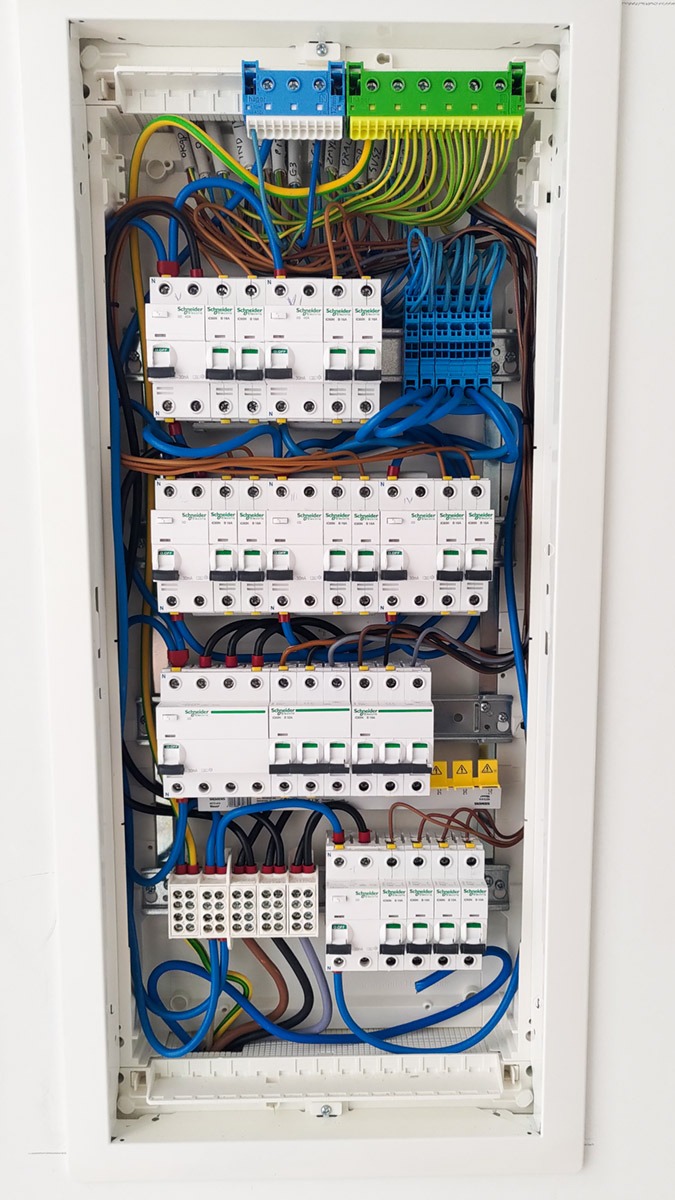 Rozdzielnica elektryczna z zainstalowanymi bezpiecznikami, listwami zaciskowymi i przewodami w różnych kolorach, umieszczona w białej obudowie.