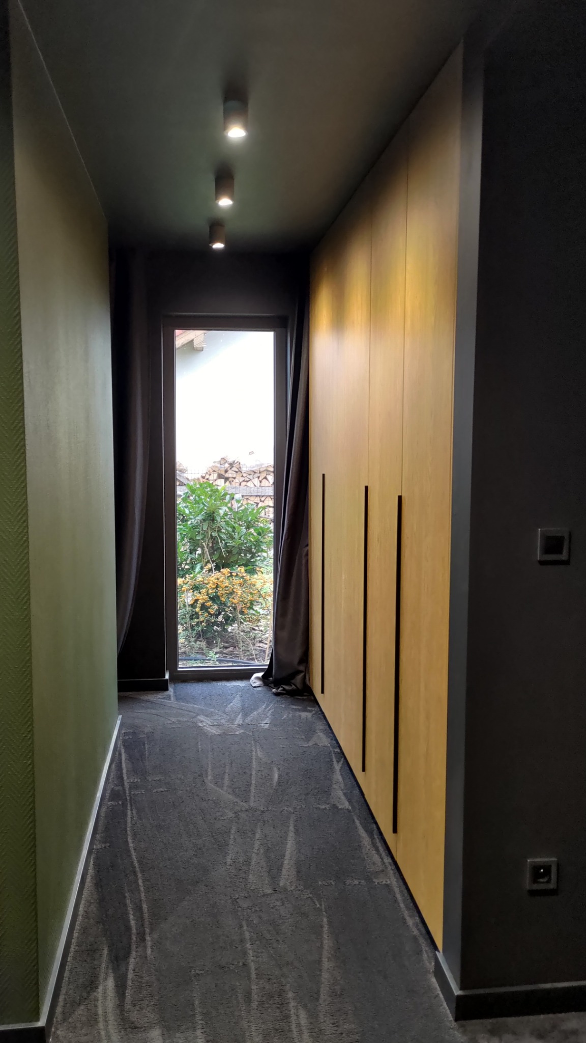 Nowoczesny korytarz z oświetleniem punktowym, drewnianą szafą w zabudowie i widokiem na ogród przez okno. Ciemna kolorystyka ścian i podłogi.