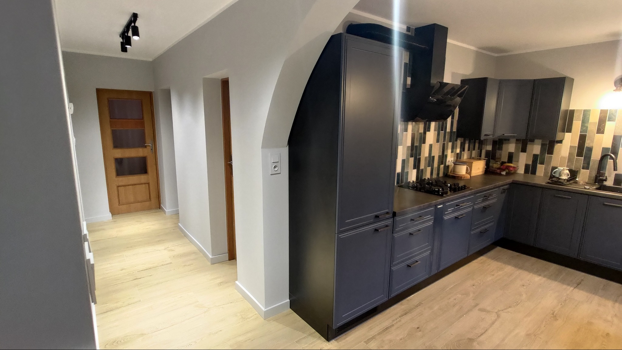 Nowoczesna, szara kuchnia z drewnianą podłogą i mozaiką na ścianie. Widoczne szafki, okap, płyta gazowa i oświetlenie sufitowe. Minimalistyczny design.