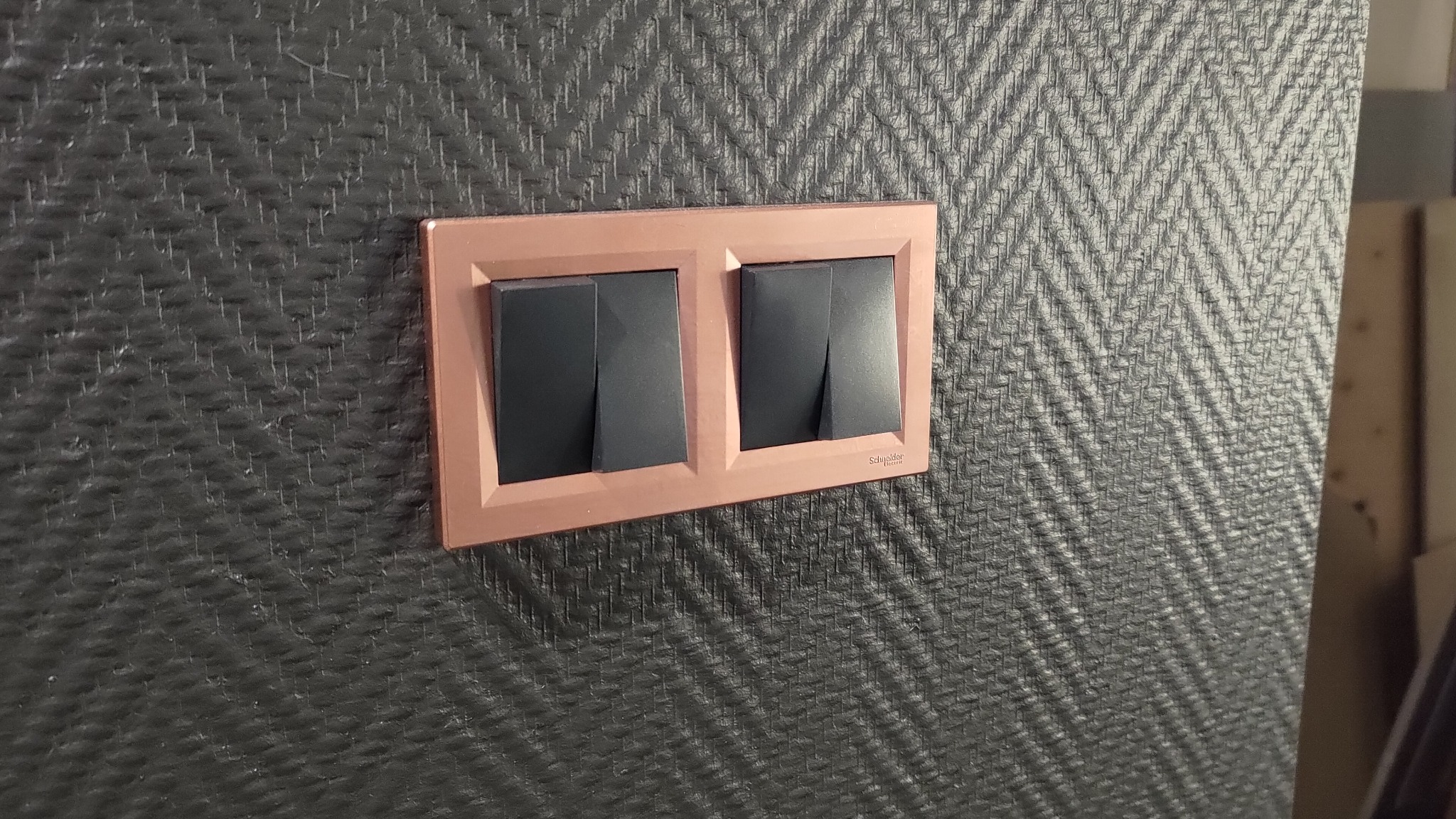 Dwa czarne włączniki światła w ramce w kolorze różowego złota, zamontowane na ścianie pokrytej tapetą w ciemny wzór jodełki. Widoczny fragment ściany i krawędź mebla.