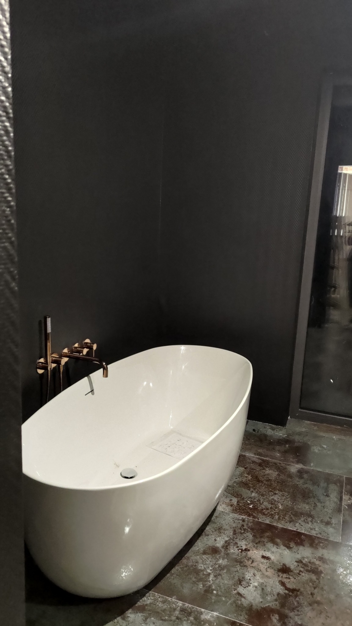 Nowoczesna, wolnostojąca wanna owalna z armaturą w kolorze różowego złota na tle ciemnych, teksturowanych ścian i podłogi z efektem rdzy. Minimalistyczny design.