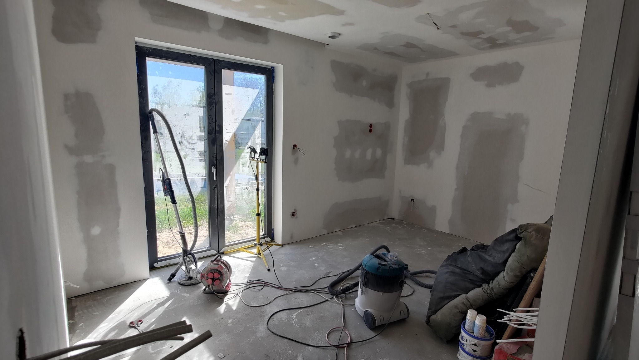 Pokój w trakcie szpachlowania ścian i sufitu, widoczne narzędzia: szlifierka, odkurzacz, lampa. Ściany częściowo pokryte gładzią, przygotowane do malowania.