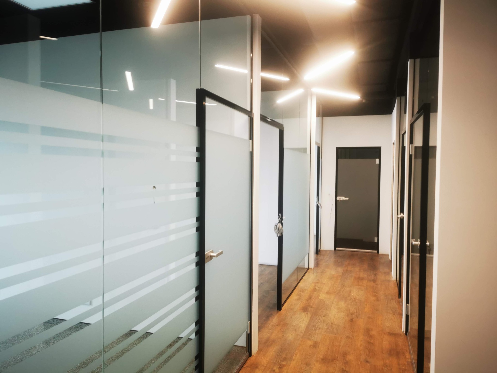 Korytarz biurowy z przeszkleniami z matowego szkła w aluminiowych ramach, podświetlony liniowymi lampami LED na czarnym suficie.
