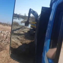 Widok w lusterku bocznym ciężarówki: koparka nabiera urobek ziemny na brzegu stawu, przygotowując teren.