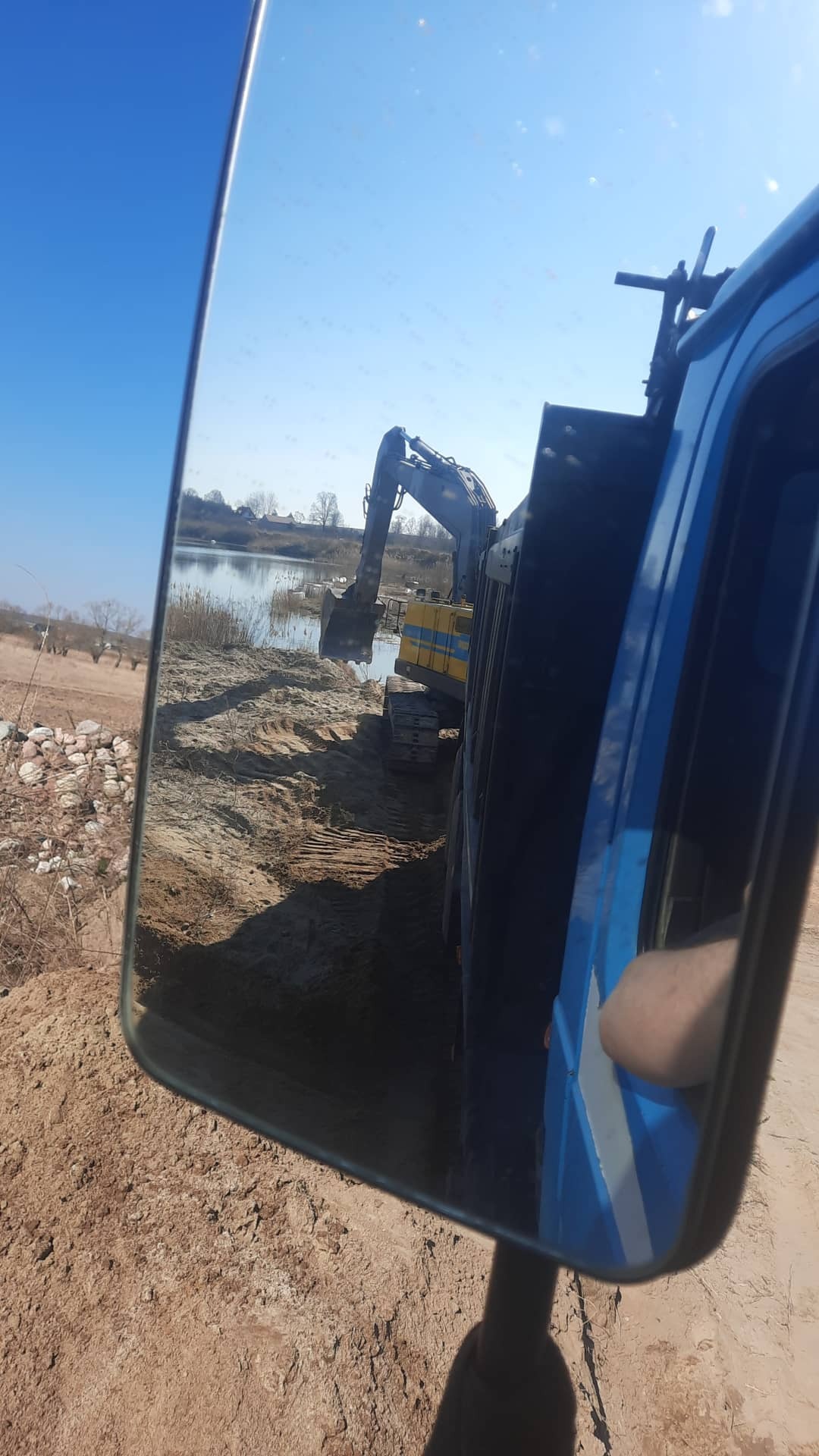 Widok w lusterku bocznym ciężarówki: koparka nabiera urobek ziemny na brzegu stawu, przygotowując teren.