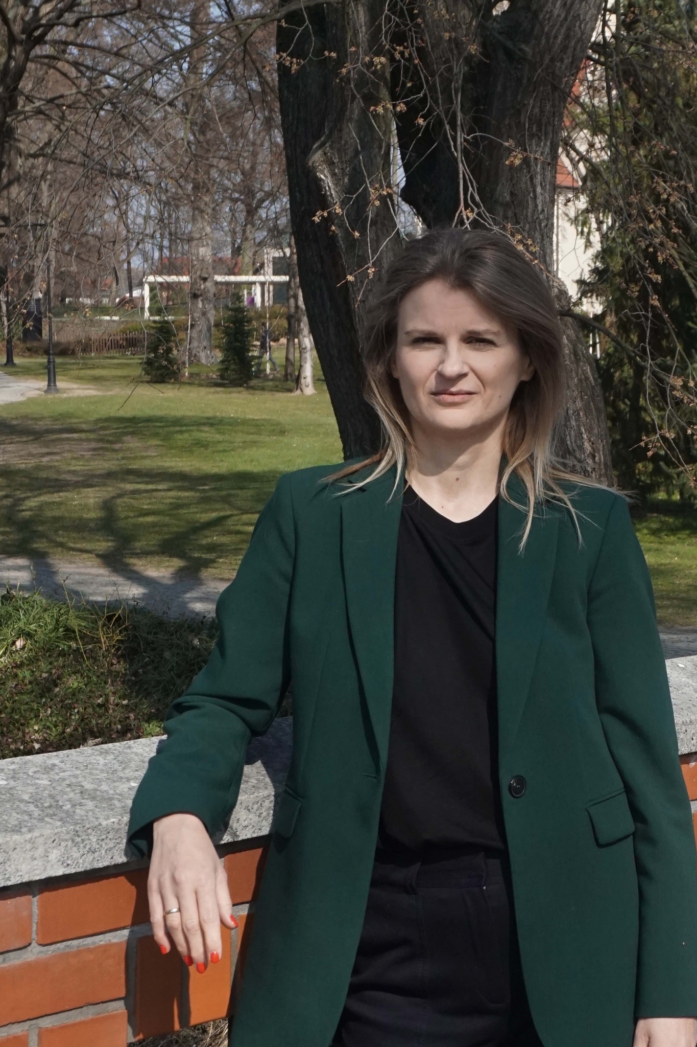 Kobieta w zielonym żakiecie opiera się o ceglany murek na tle parku z drzewami bez liści i budynkami w oddali.