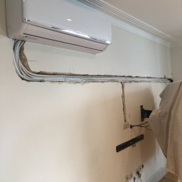 Remontu i tam - Biały klimatyzator ścienny zamontowany nad wyciętym korytarzem w ścianie, przez który prowadzone są przewody. Widoczne gniazdka elektryczne i uchwyt do telewizora w trakcie instalacji.