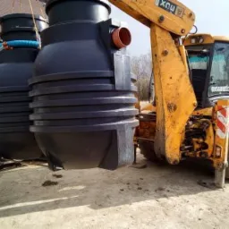 Żółta koparko-ładowarka transportuje czarny, plastikowy zbiornik na ścieki z pomarańczową rurą wlotową, widoczne trzy podobne zbiorniki w tle.