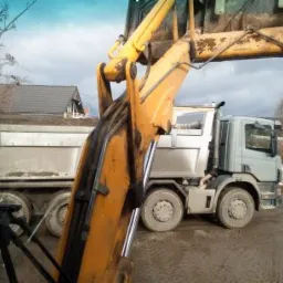 Żółta koparka załadowuje ziemię do szarej ciężarówki na placu budowy, w tle dom z ciemnym dachem i osoba w żółtej kurtce.