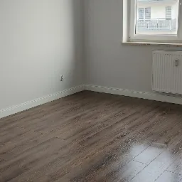 Pokój z ułożonymi ciemnymi panelami podłogowymi, świeżo pomalowanymi ścianami na biało i widokiem z okna na sąsiedni budynek.