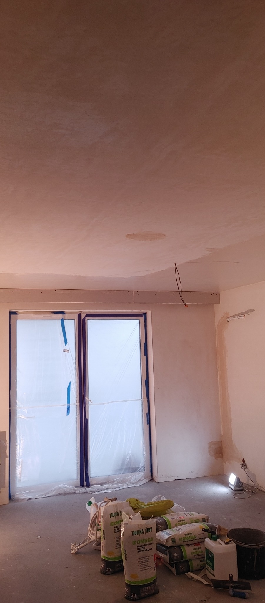Wnętrze w trakcie prac wykończeniowych: okno zabezpieczone folią, worki z gładzią gipsową Dolina Nidy na podłodze, surowy sufit z widocznymi śladami po łataniu, punktowe oświetlenie robocze.