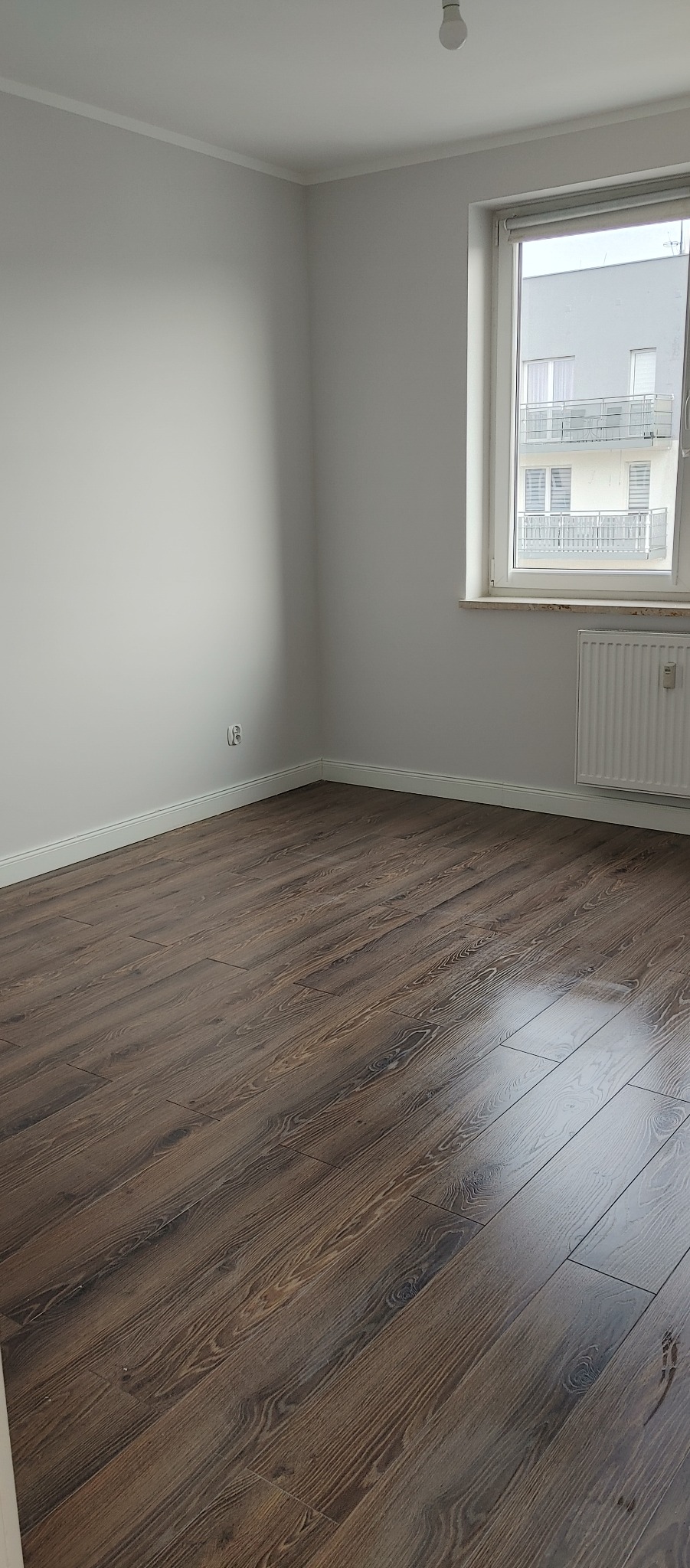 Pokój z ułożonymi ciemnymi panelami podłogowymi, świeżo pomalowanymi ścianami na biało i widokiem z okna na sąsiedni budynek.