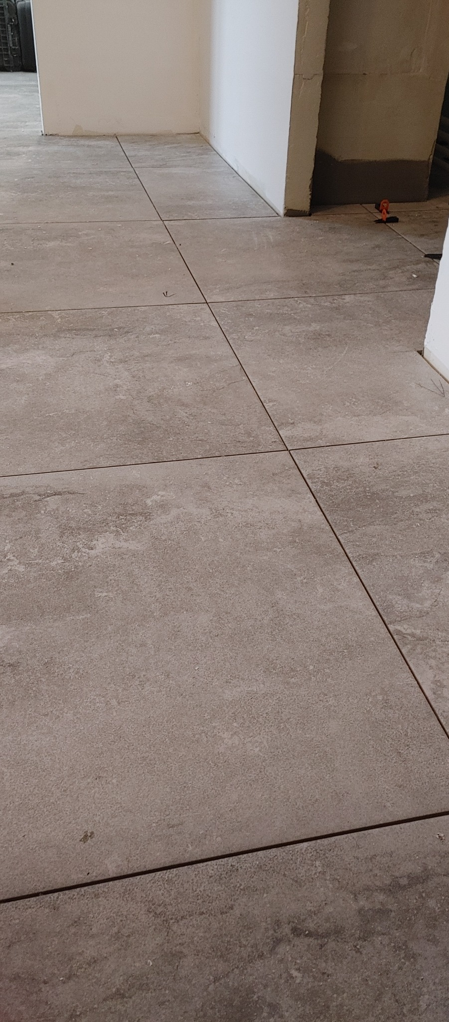 Ułożone, beżowe płytki podłogowe z ciemnymi fugami, widoczne fragmenty ścian w surowym stanie.