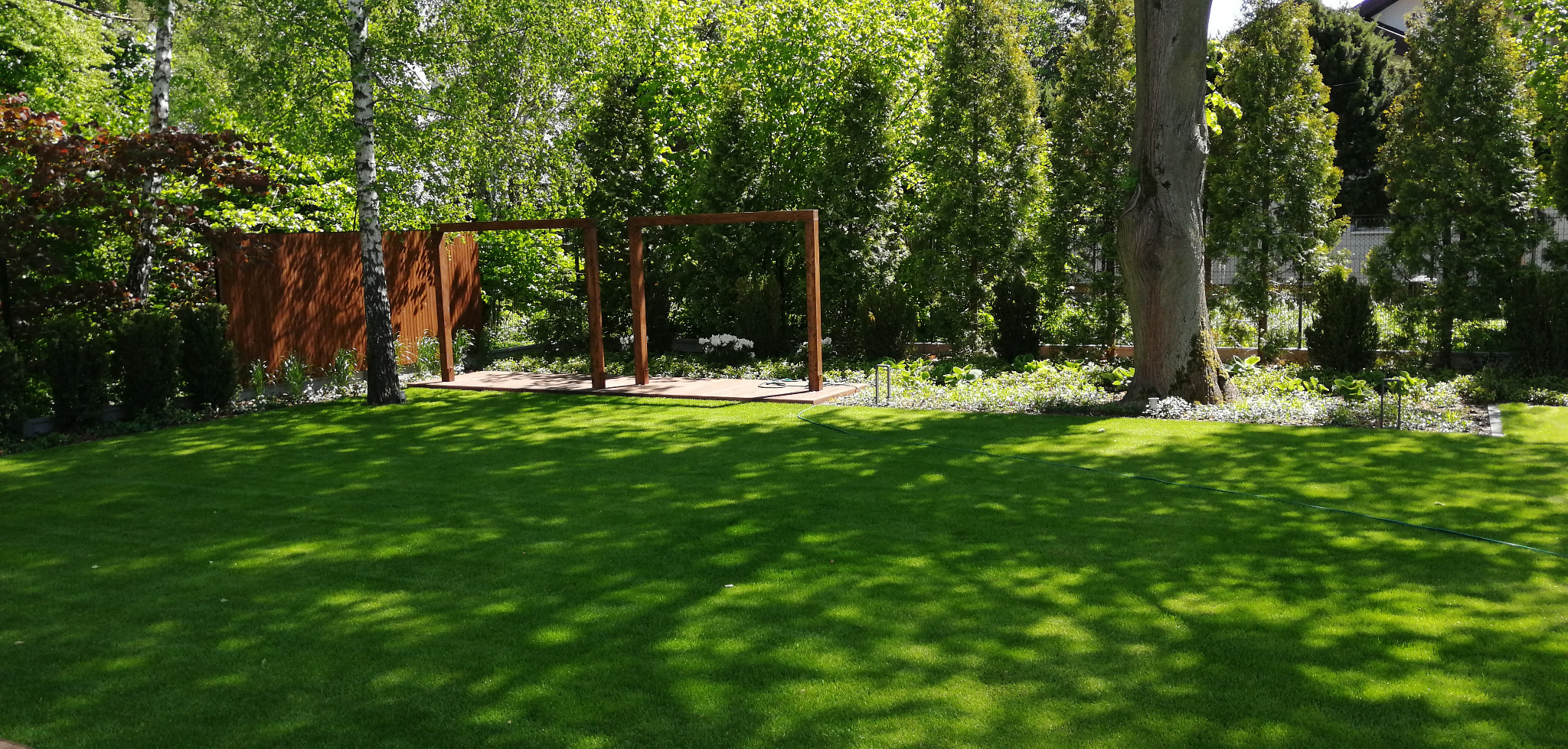 Zadbany ogród z idealnie przystrzyżonym trawnikiem, drewnianą pergolą i elementami dekoracyjnymi, widok na zielone drzewa i krzewy w tle.