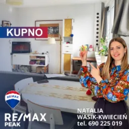 Agentka nieruchomości w kwiecistej bluzce prezentuje jasne mieszkanie z okrągłym stołem, telewizorem i wysoką szafką, wskazując na przestrzeń; widoczne logo agencji RE/MAX i dane kontaktowe.