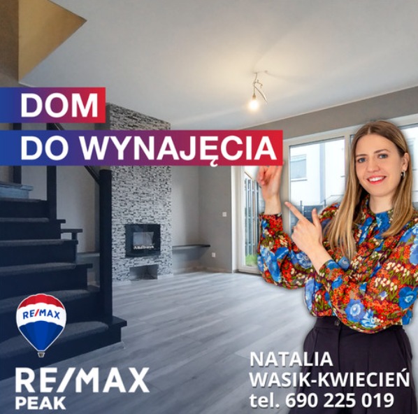 Wnętrze domu z kominkiem i schodami, z napisem 'Dom do wynajęcia' i agentką nieruchomości w kwiecistej bluzce wskazującą na przestrzeń, logo RE/MAX Peak i danymi kontaktowymi Natalii Wasik-Kwiecień.