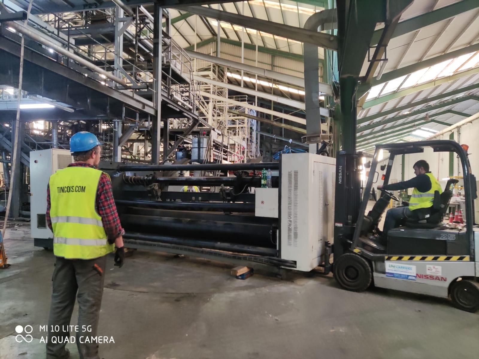 Pracownik w kasku i kamizelce odblaskowej obserwuje maszynę przemysłową, w tle wózek widłowy i konstrukcja hali.