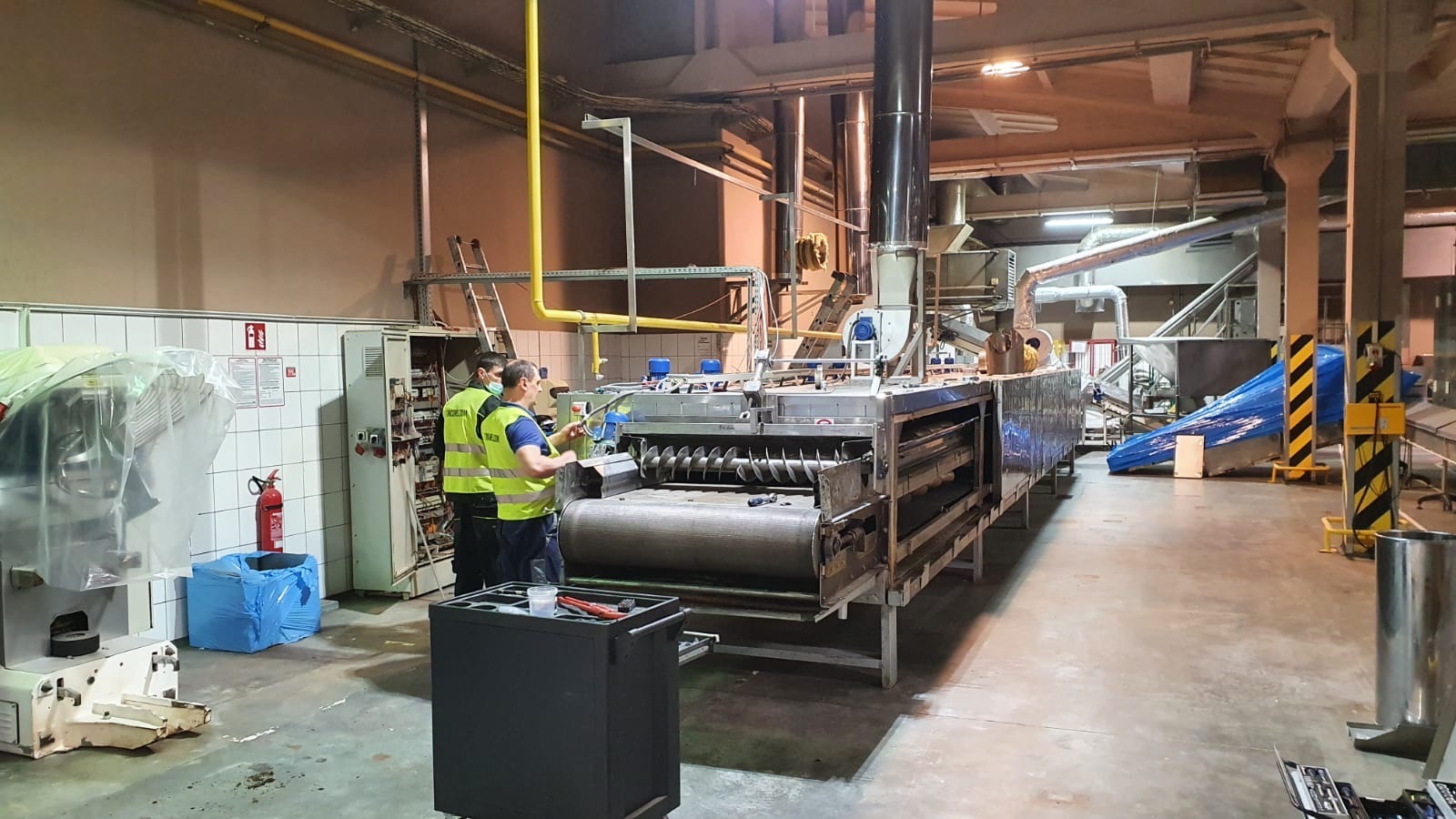 Dwóch serwisantów w kamizelkach, z narzędziami, dokonuje przeglądu i naprawy dużej, przemysłowej maszyny z elementami spiralnymi w hali produkcyjnej, widoczne drabiny i instalacje pod sufitem.