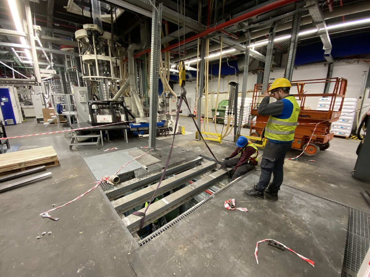 Dwóch pracowników w kaskach, jeden stojący w kamizelce odblaskowej, drugi klęczący, pracują przy otworze w podłodze w hali przemysłowej z widoczną maszyną do przetwórstwa tworzyw sztucznych w tle.