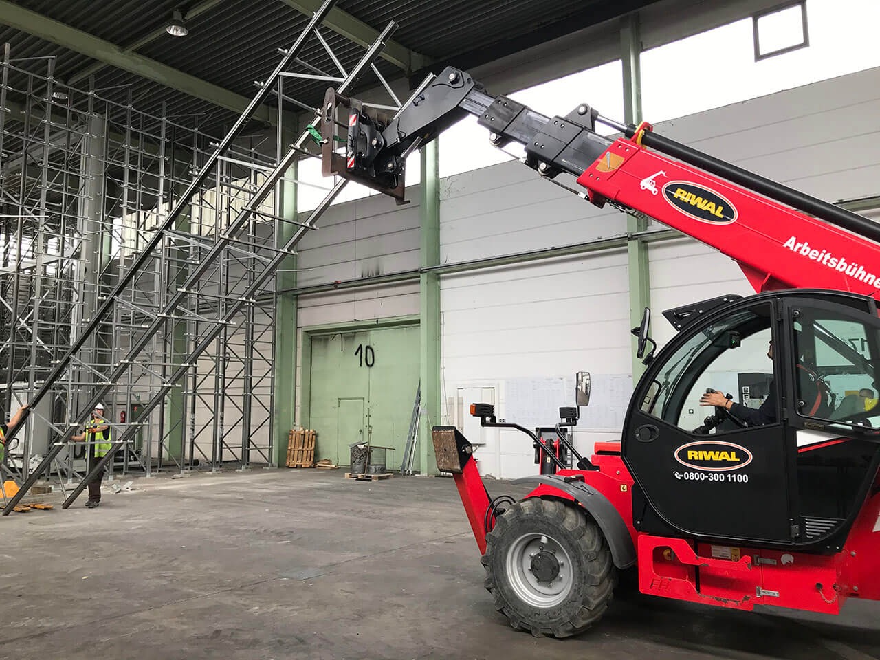 Czerwony wózek widłowy teleskopowy marki Riwal podnosi metalową konstrukcję w hali przemysłowej, pracownik w kamizelce odblaskowej nadzoruje proces.